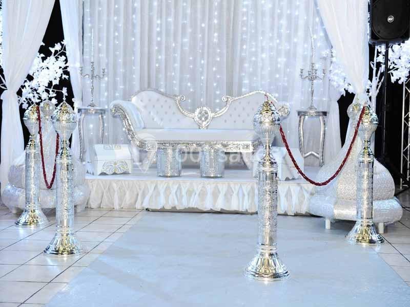 Décor de mariage élégant avec canapé blanc orné, rideaux lumineux et poteaux argentés reliés par des cordons rouges.