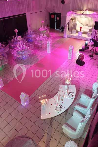 Salle de réception décorée avec des tables, chaises transparentes, tapis rose et une scène ornée d’un canapé blanc.