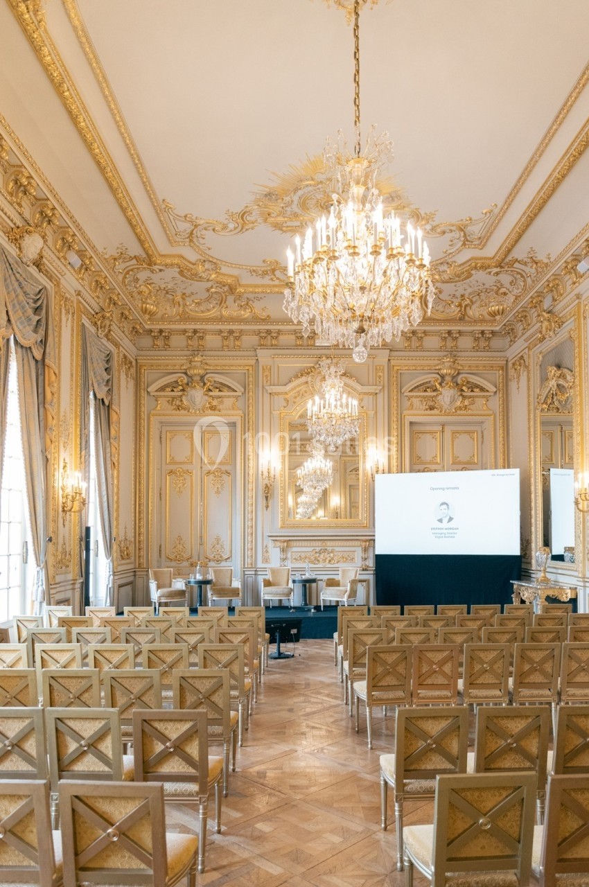 Salle de conférence élégante avec décorations dorées, chandeliers et rangées de chaises alignées devant un écran.