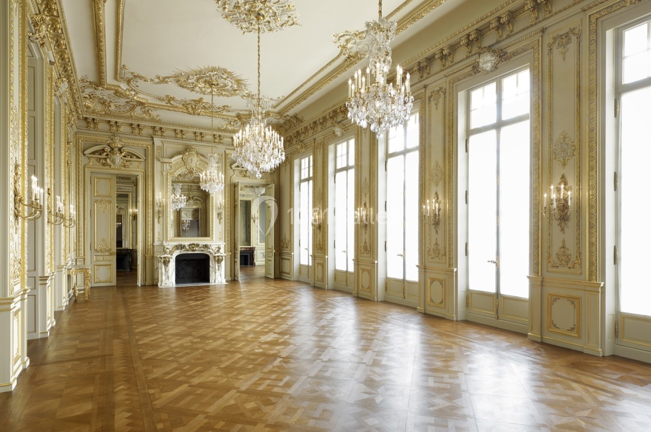 Salle de réception élégante avec parquet en bois, moulures dorées, lustres en cristal et grandes fenêtres.