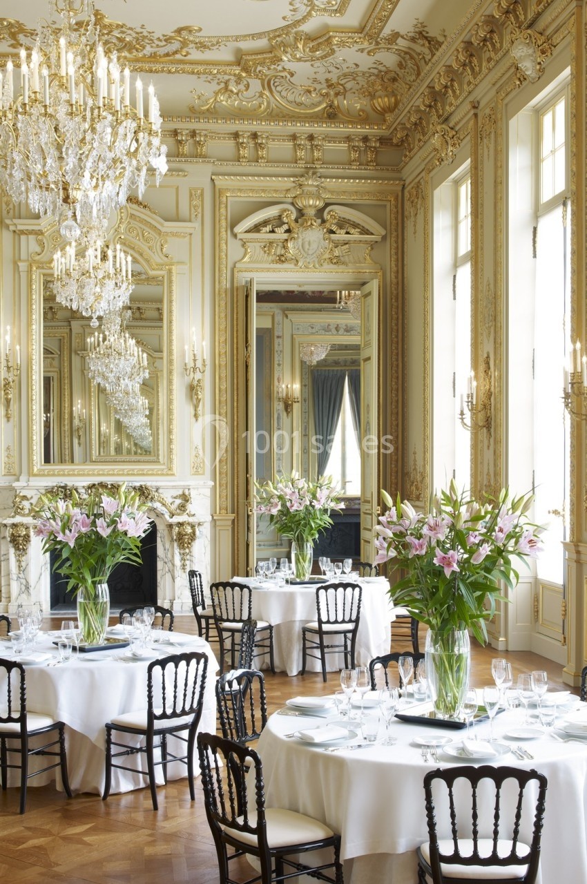 Salle de réception élégante avec moulures dorées, lustres en cristal et tables dressées avec nappes blanches et fleurs.