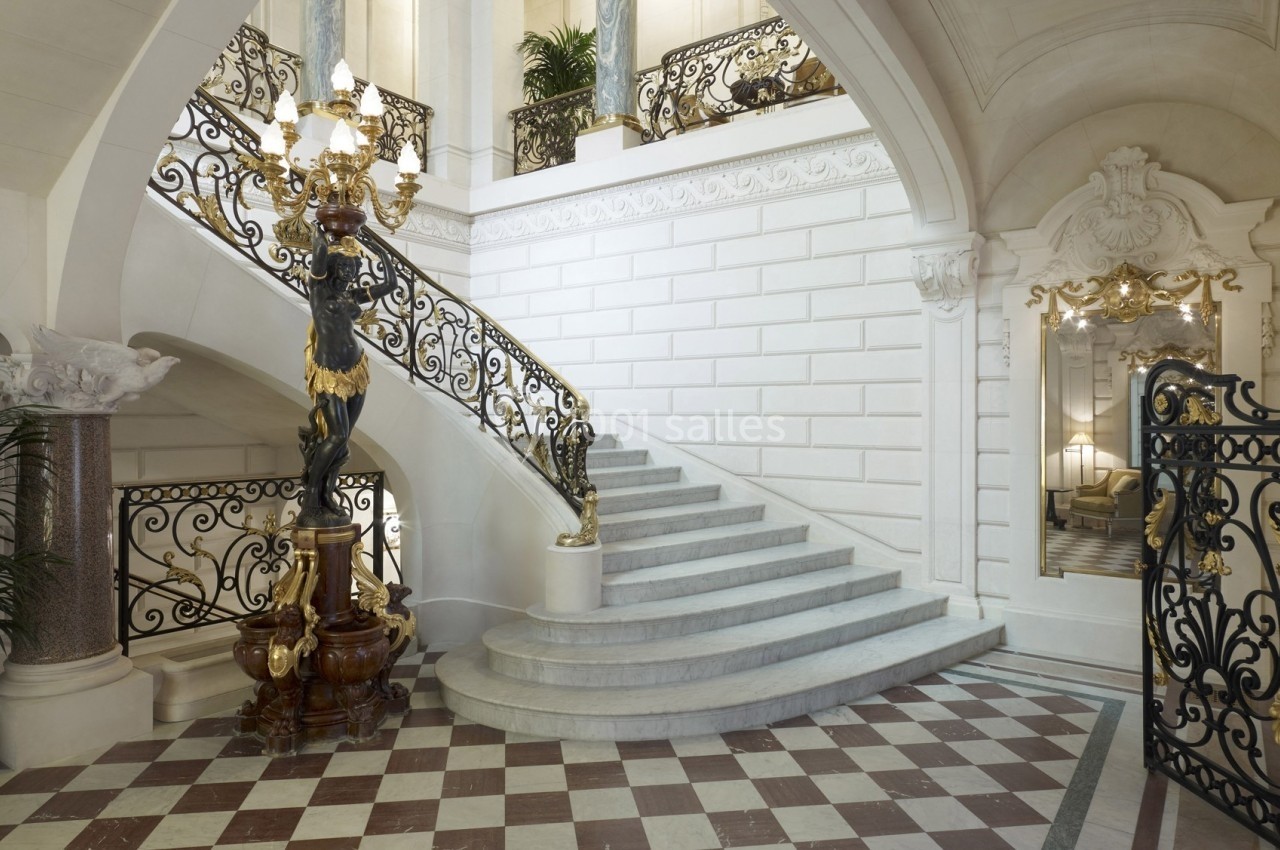 Escalier en marbre blanc avec rampe en fer forgé orné, dans un intérieur élégant avec sol en damier et lustre décoratif.