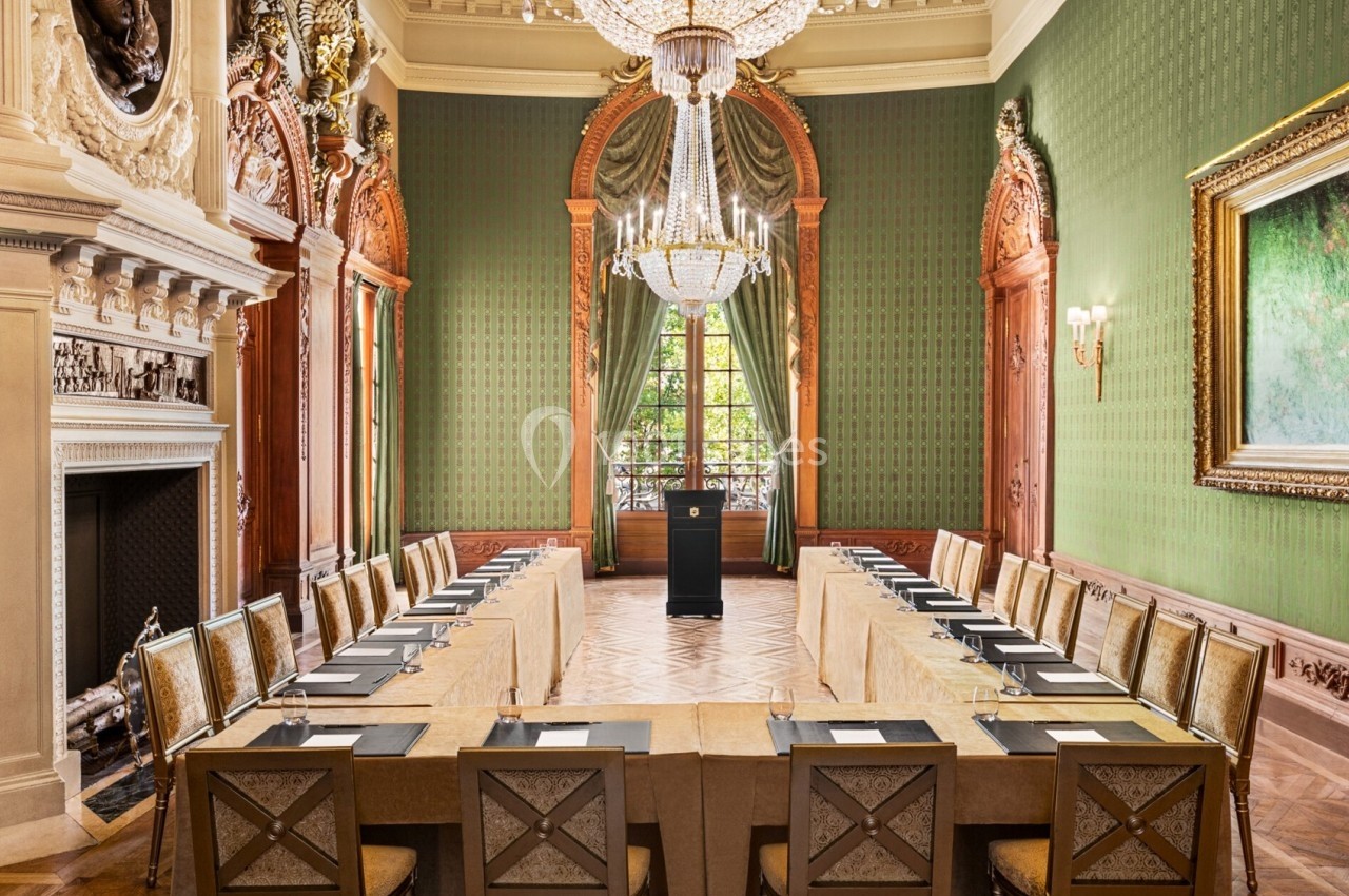Salle de réunion élégante avec une table en U, chaises dorées, lustres, murs verts et grandes fenêtres donnant sur un jardin.