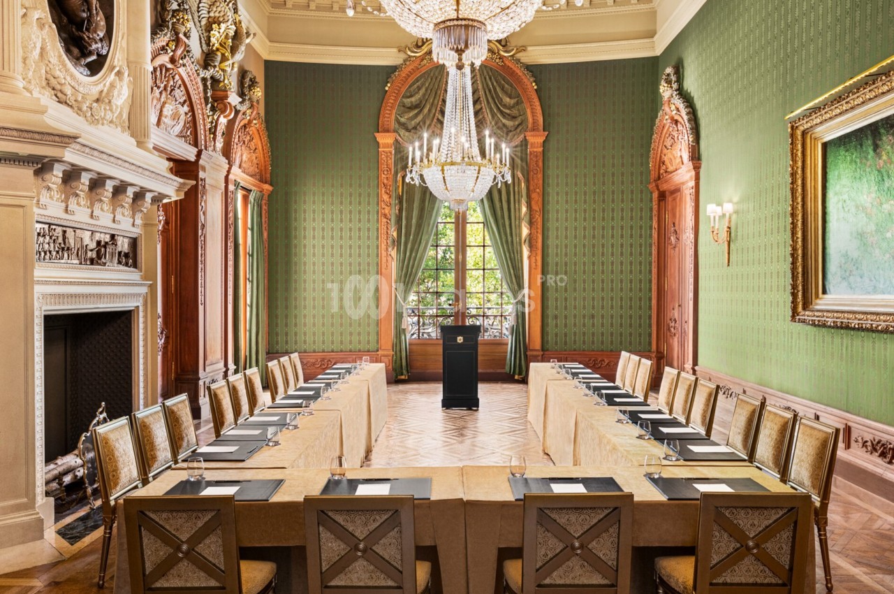 Salle de réunion élégante avec une table en U, chaises dorées, lustres, murs verts et grandes fenêtres donnant sur un jardin.
