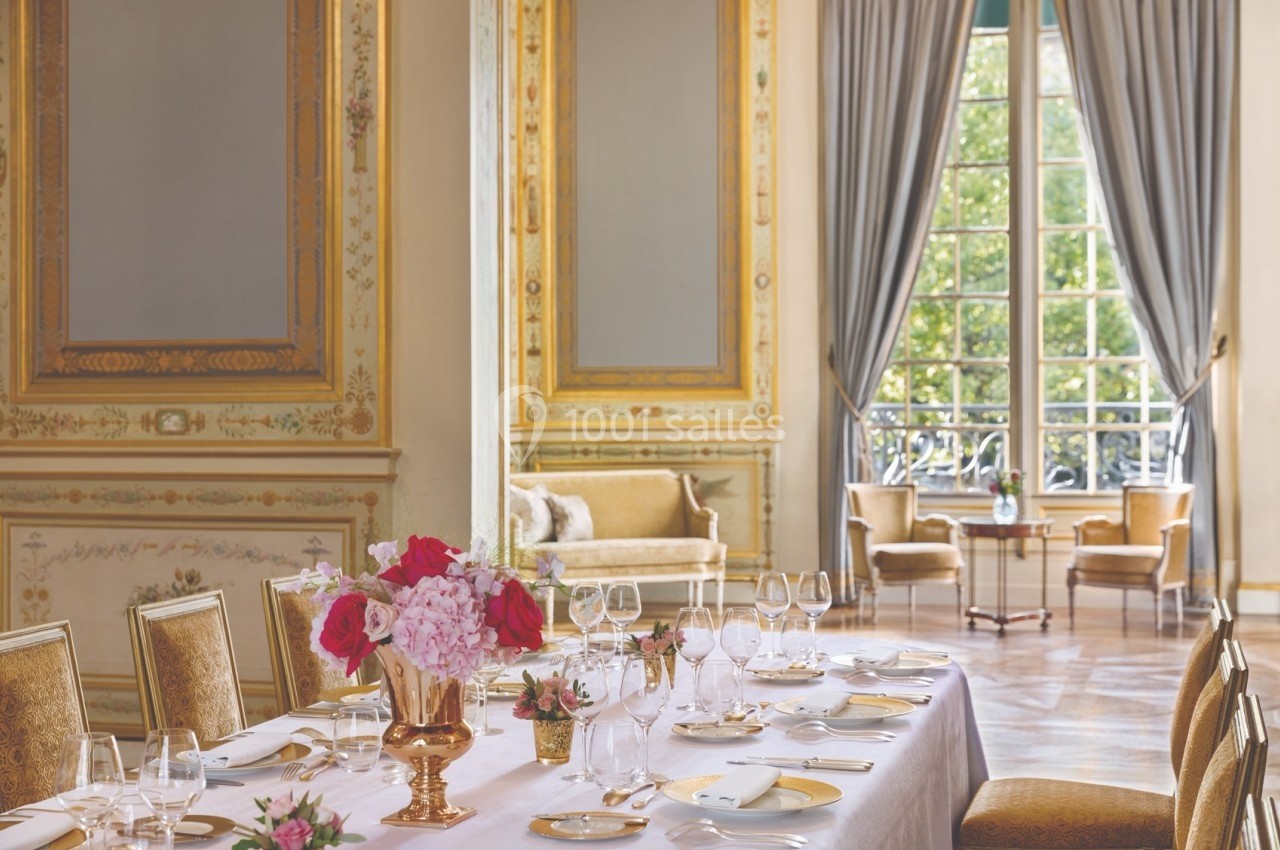 Salle élégante avec table dressée, décorée de fleurs, moulures dorées, grandes fenêtres et rideaux ouverts sur un jardin.