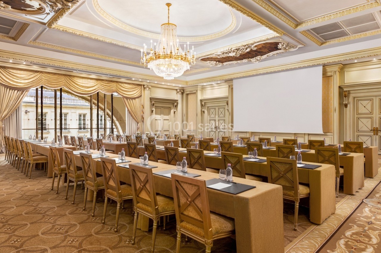 Salle de conférence élégante avec des rangées de tables et chaises, éclairée par un grand lustre et une lumière naturelle.