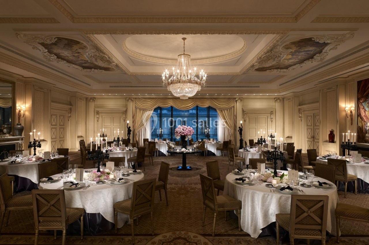 Salle de réception élégante avec tables rondes dressées, lustre central et grandes fenêtres donnant sur l'extérieur.