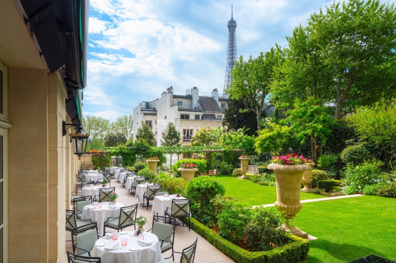 Terrasse d'un restaurant élégant avec des tables dressées, vue sur un jardin verdoyant et la tour Eiffel en arrière-plan.