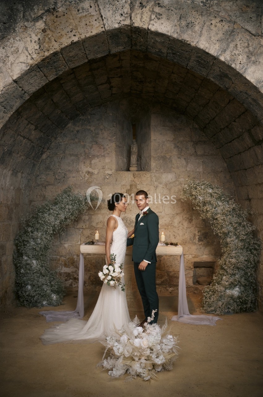 Un couple en tenue de mariage pose devant un autel décoré de fleurs dans une chapelle en pierre.