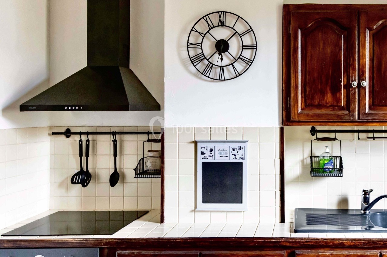 Cuisine avec hotte noire, plaques de cuisson, évier, horloge murale et rangements suspendus sur carrelage blanc.