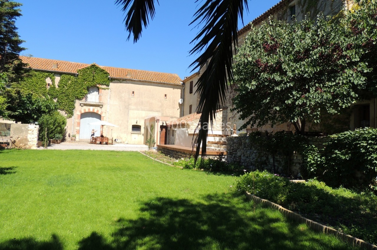 Cour verdoyante entourée de bâtiments anciens en pierre, avec des arbres et un ciel bleu dégagé.
