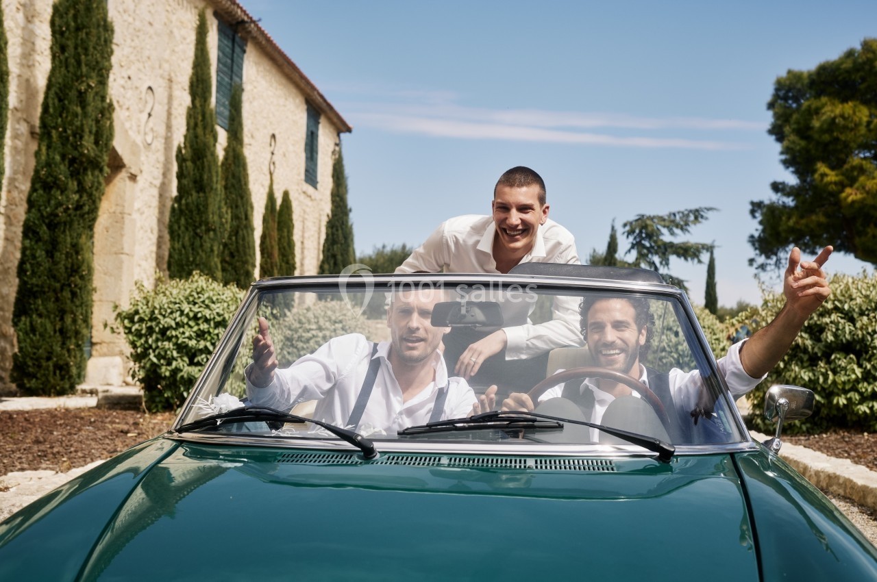 Trois hommes souriants dans une voiture décapotable verte devant un bâtiment en pierre et des arbres.