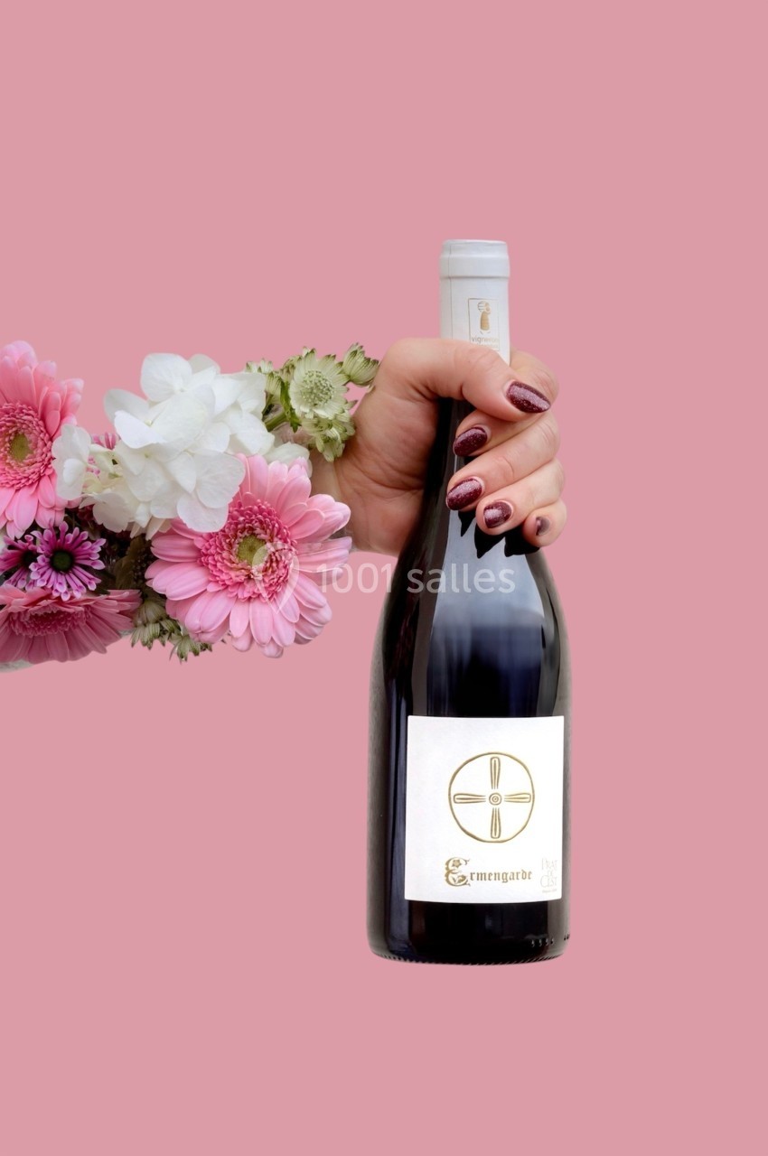 Une main tenant une bouteille de vin avec une étiquette minimaliste, entourée de fleurs colorées sur fond rose.