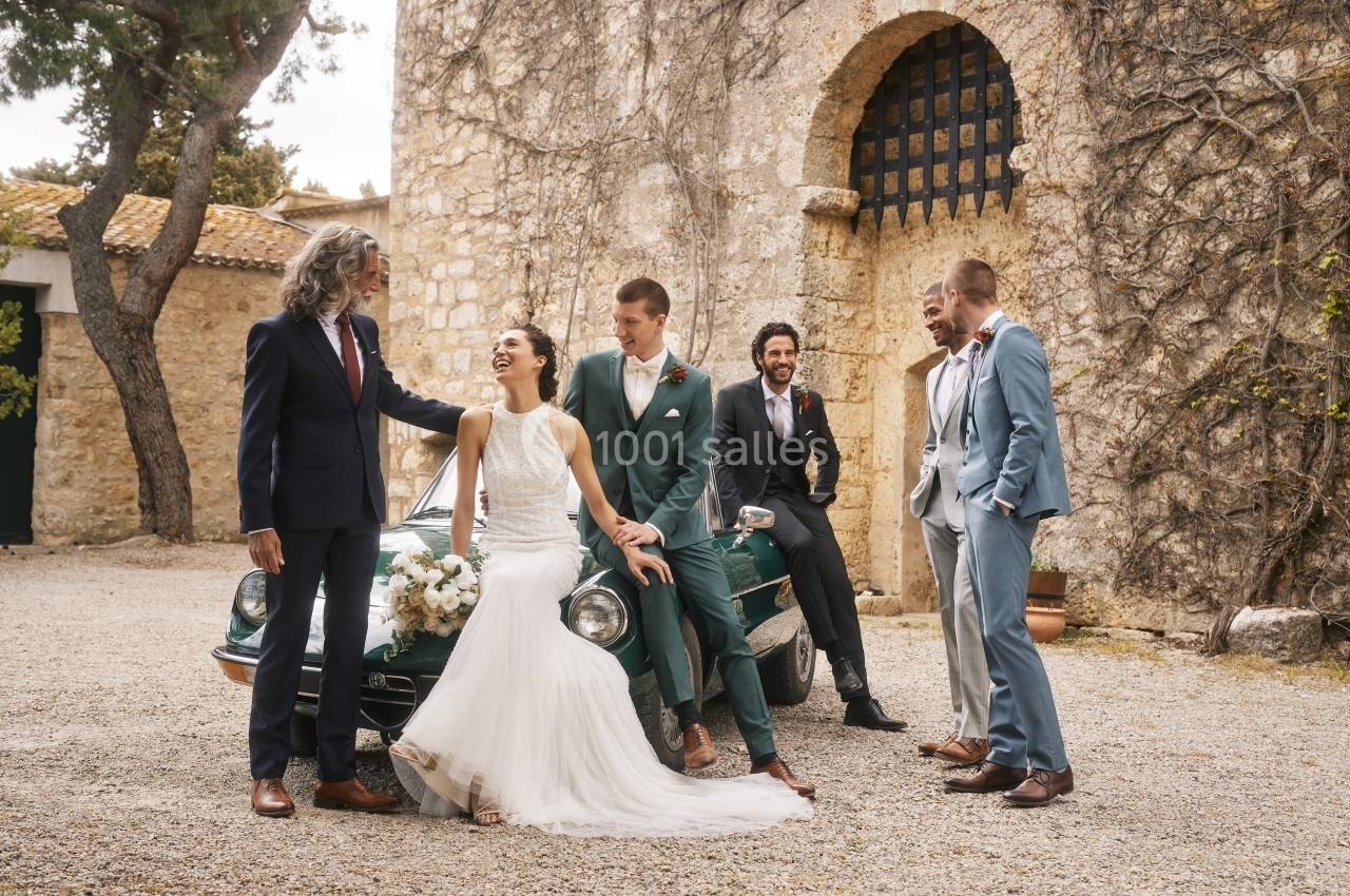 Un couple de mariés pose avec des invités élégants devant une voiture ancienne et un bâtiment en pierre.