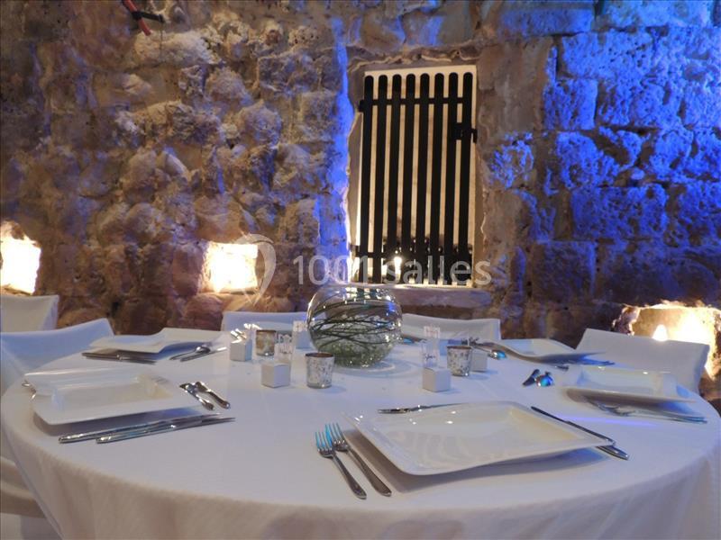 Table ronde dressée avec une nappe blanche, vaisselle moderne et un centre de table en verre, dans une salle en pierre…
