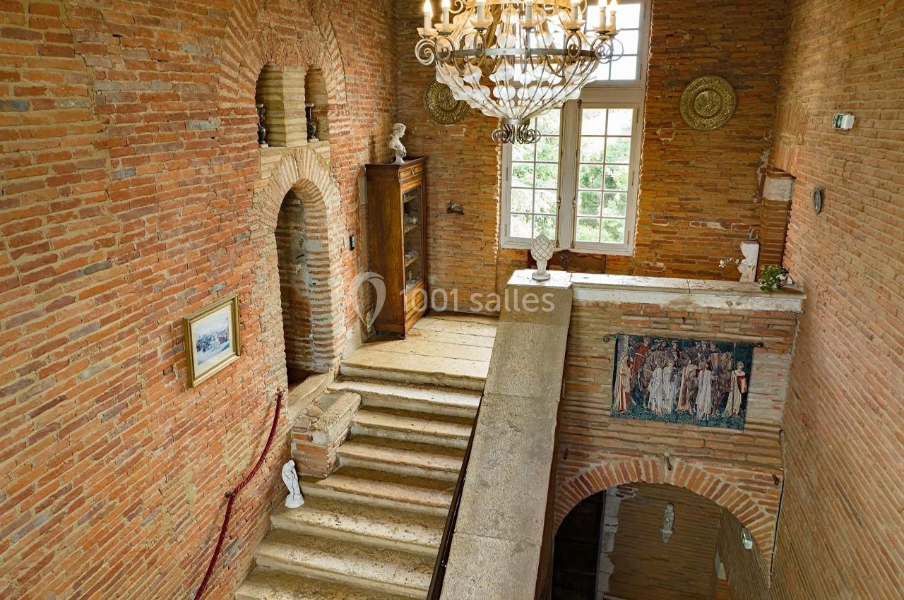 Escalier en pierre dans un intérieur en briques rouges, éclairé par un lustre et une fenêtre avec vue sur la verdure.