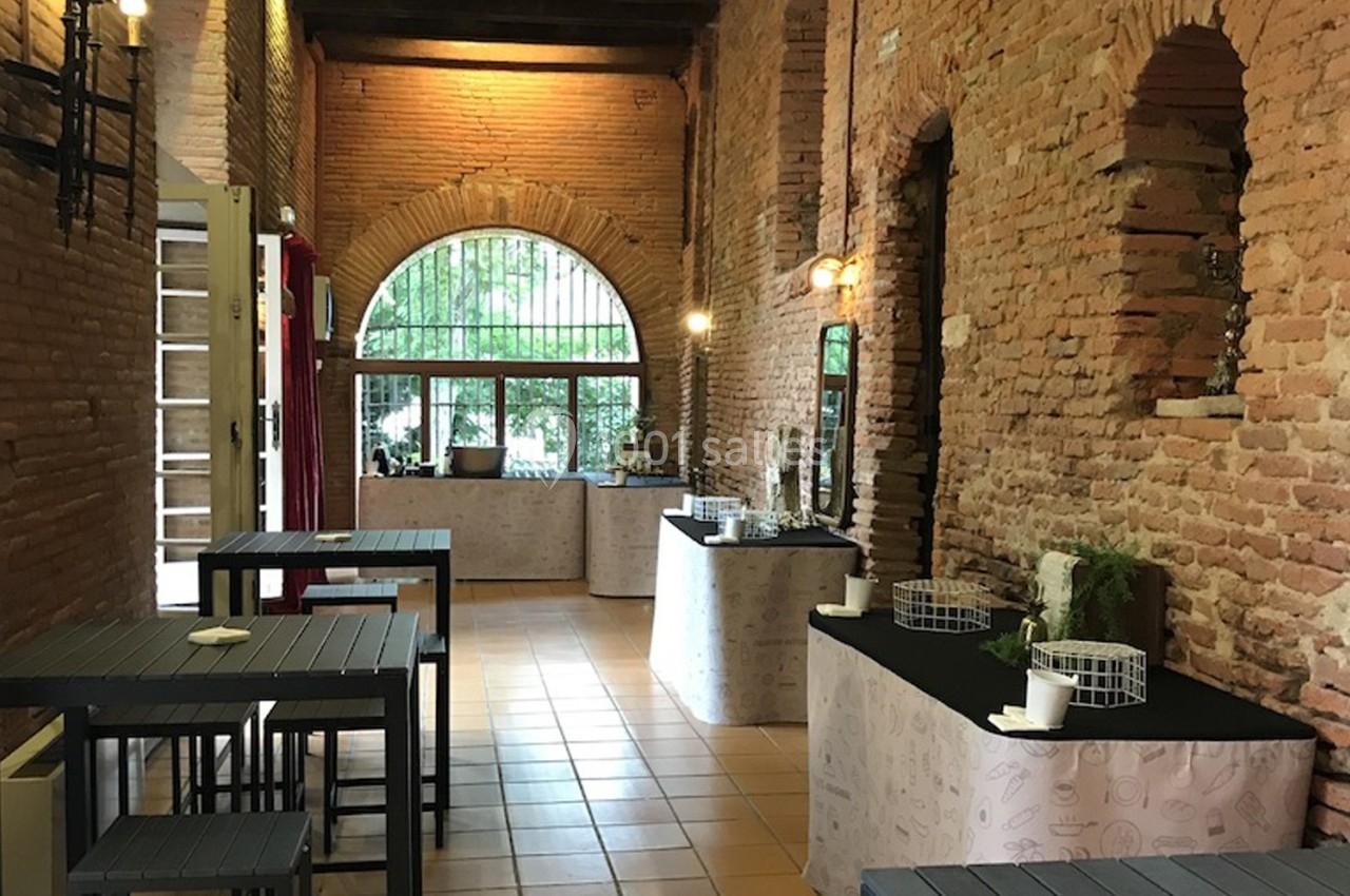 Salle en briques avec tables hautes noires, buffets décorés et grande fenêtre en arche donnant sur un espace vert.