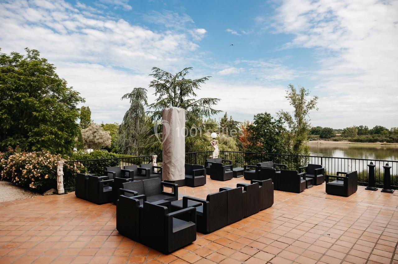 Terrasse en briques avec des fauteuils noirs, entourée de végétation et offrant une vue sur un plan d'eau.