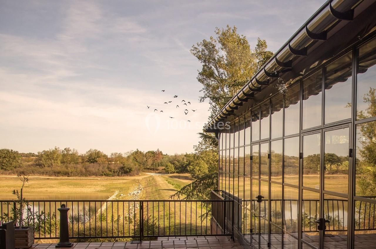 Terrasse en carrelage avec garde-corps, vue sur un paysage naturel avec arbres, champs et un groupe d'oiseaux en vol.