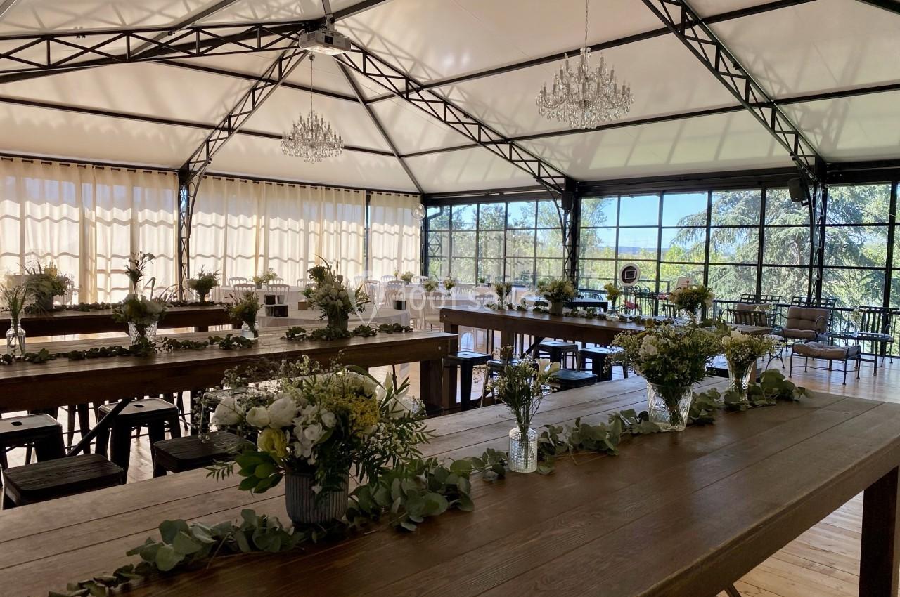 Salle lumineuse avec de grandes baies vitrées, tables en bois décorées de fleurs et guirlandes végétales.