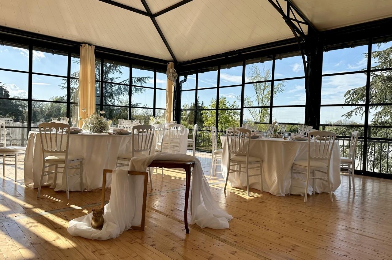 Salle lumineuse avec grandes baies vitrées, tables dressées avec nappes blanches et décorations élégantes.