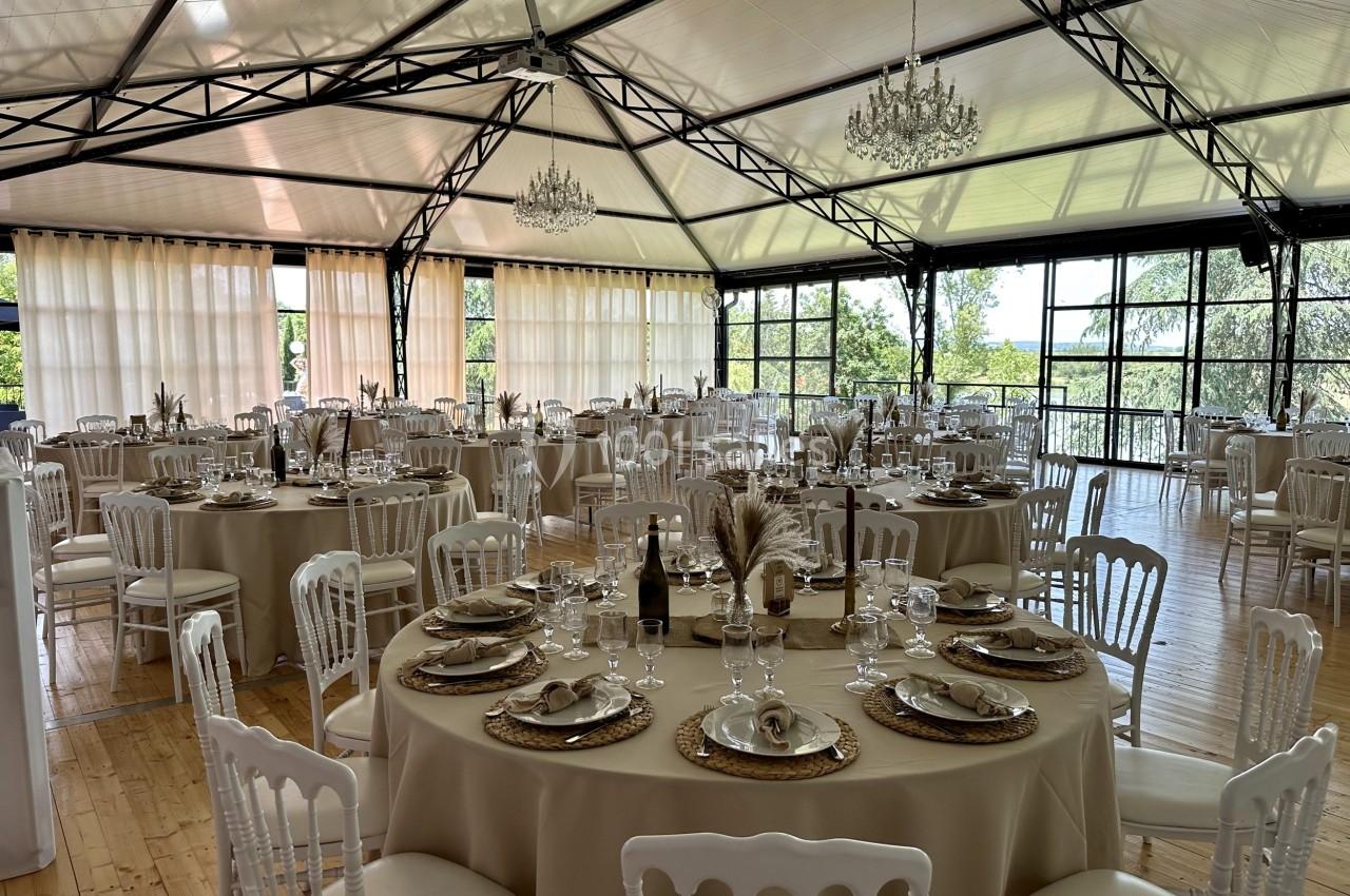 Salle de réception lumineuse avec tables rondes dressées, nappes beiges, chaises blanches et grandes baies vitrées.