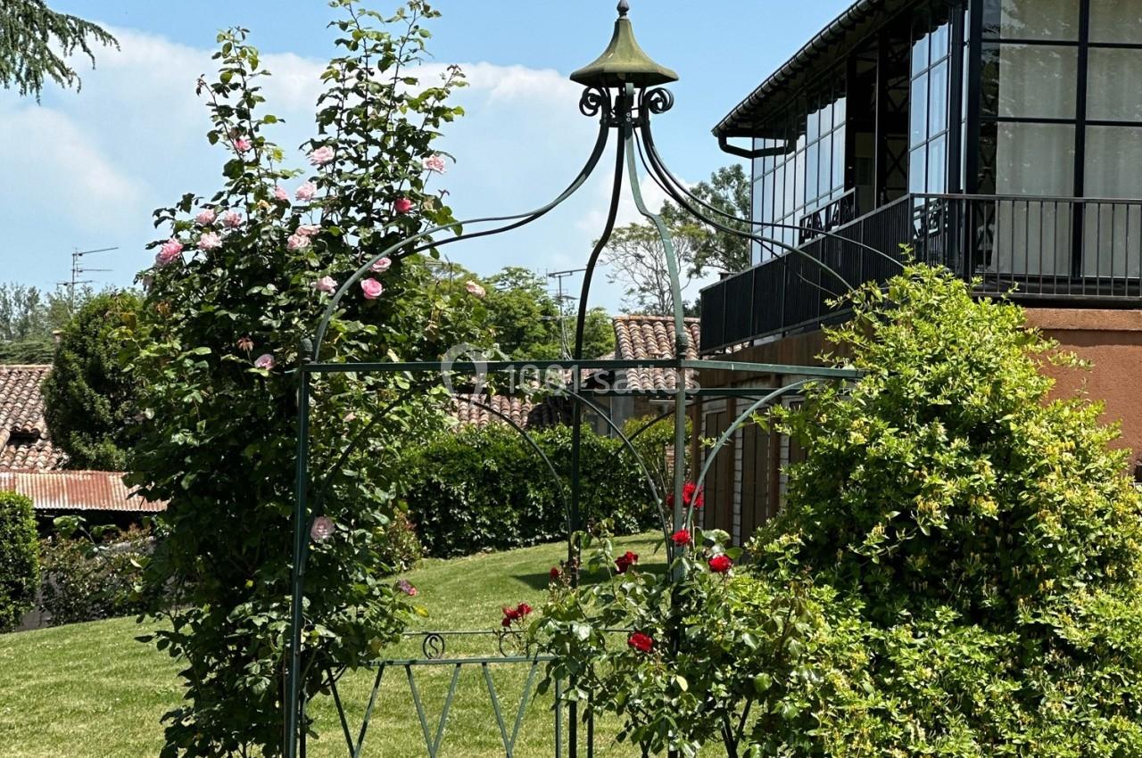 Arche en métal ornée de roses grimpantes dans un jardin verdoyant, près d'une maison avec une véranda vitrée.