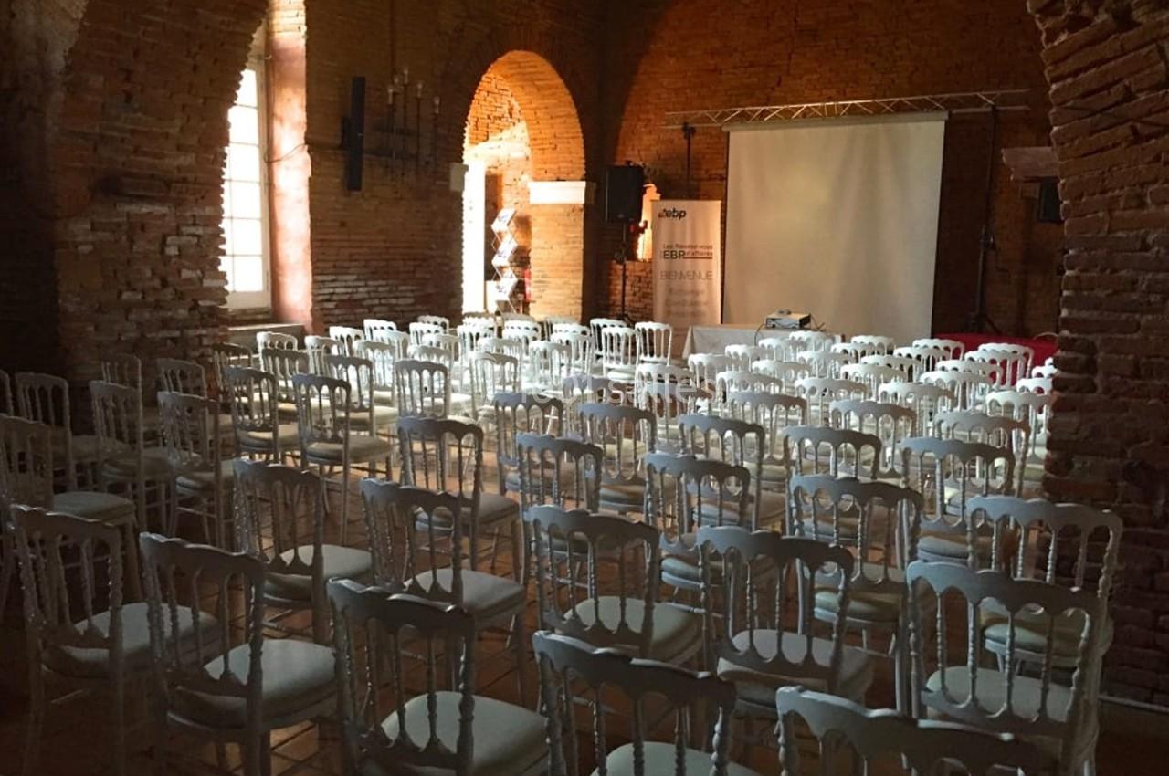 Salle en briques avec des rangées de chaises blanches disposées face à un écran de projection.
