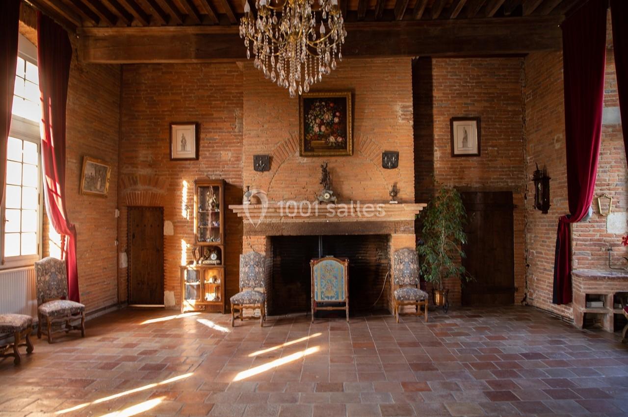 Salle avec murs en briques, cheminée centrale, lustre en cristal, meubles anciens et grandes fenêtres avec rideaux rouges.