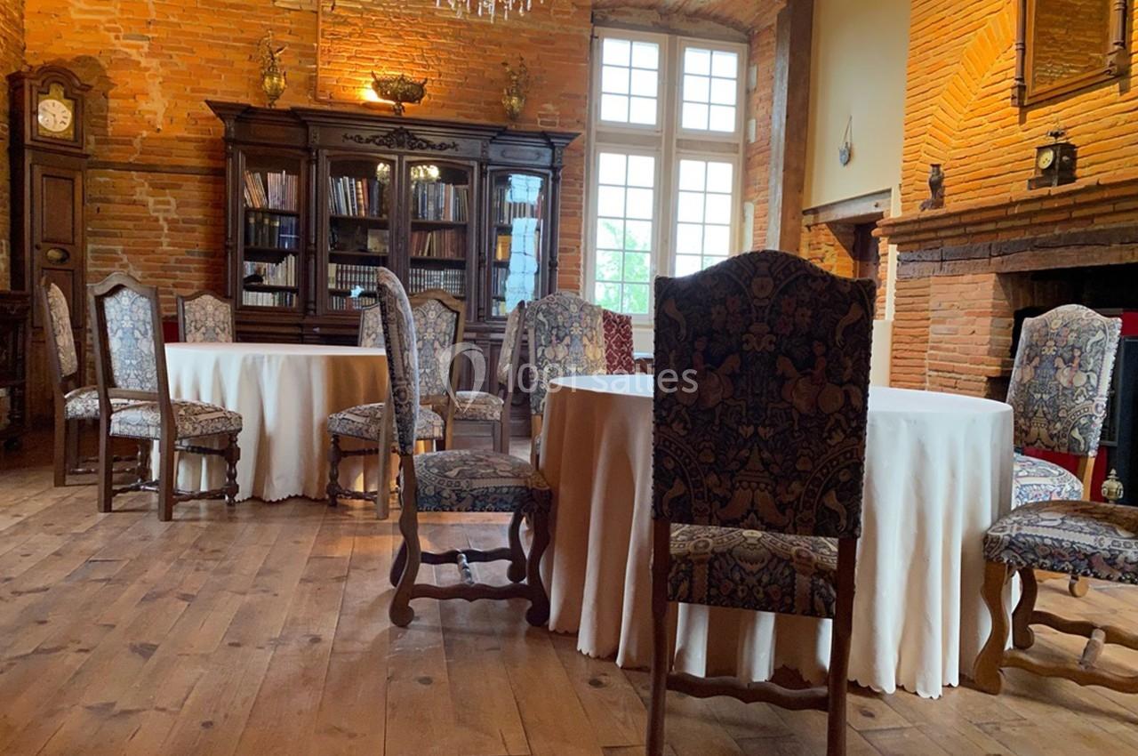 Salle avec des tables rondes couvertes de nappes blanches, chaises en bois sculpté et murs en briques apparentes.