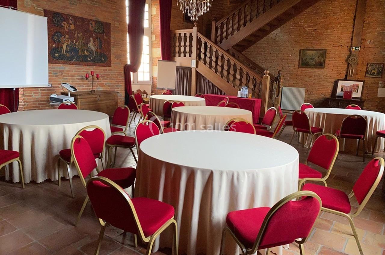 Salle avec des tables rondes nappées de blanc, entourées de chaises rouges, décorée de murs en briques et d'un lustre.