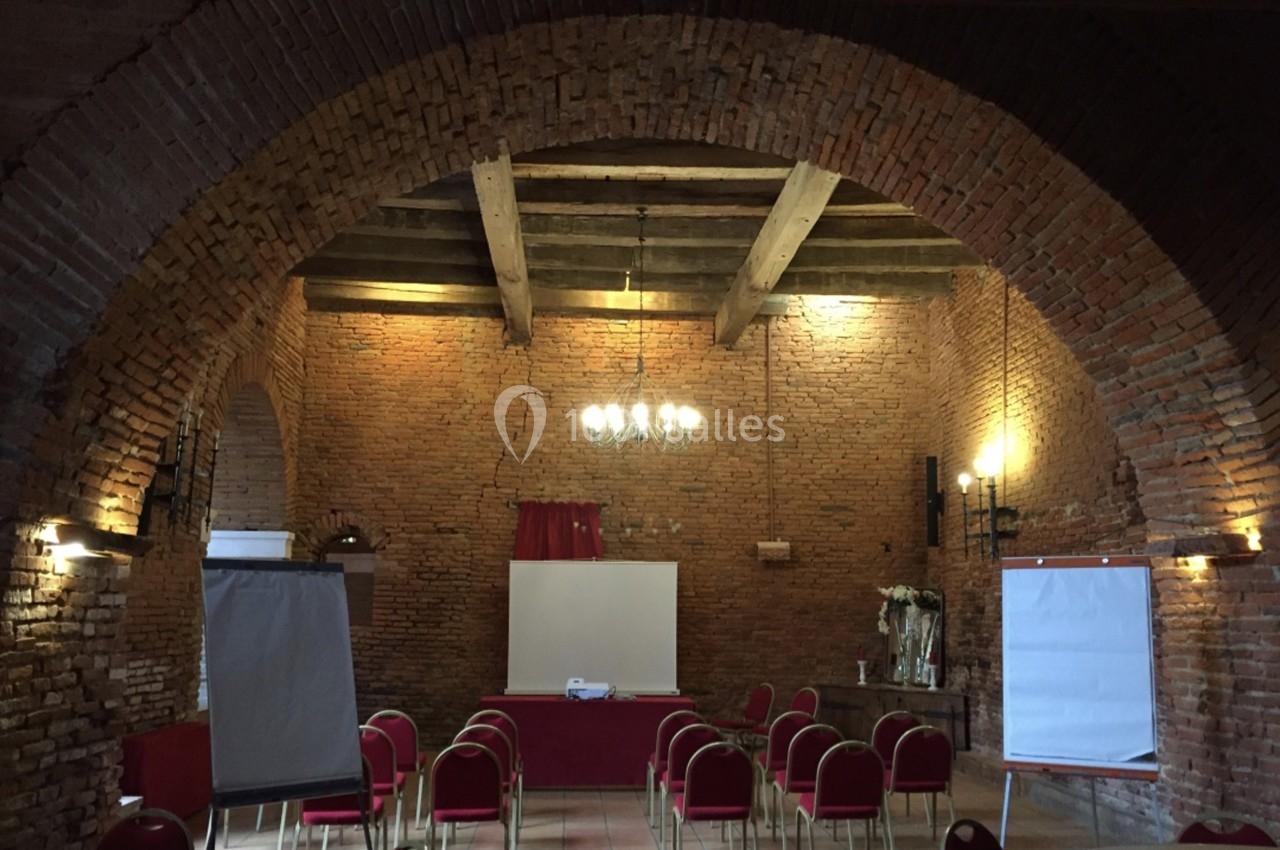 Salle en briques avec des voûtes, des chaises rouges disposées face à un écran de projection et des chevalets.
