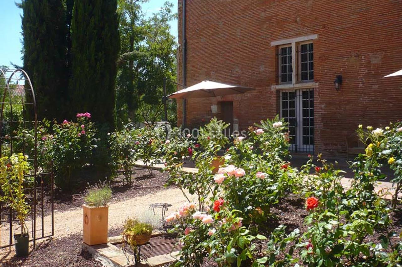 Jardin fleuri avec rosiers, allées en gravier, parasols et façade en briques rouges en arrière-plan.