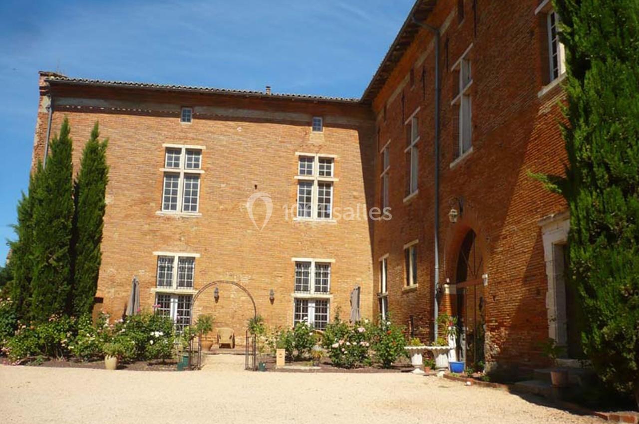Façade en briques d'un bâtiment ancien avec fenêtres à croisillons, entouré de végétation et d'une cour en gravier.