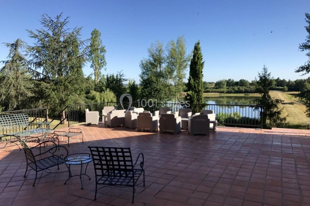 Terrasse en carrelage rouge avec mobilier de jardin, vue sur un étang entouré d'arbres sous un ciel dégagé.