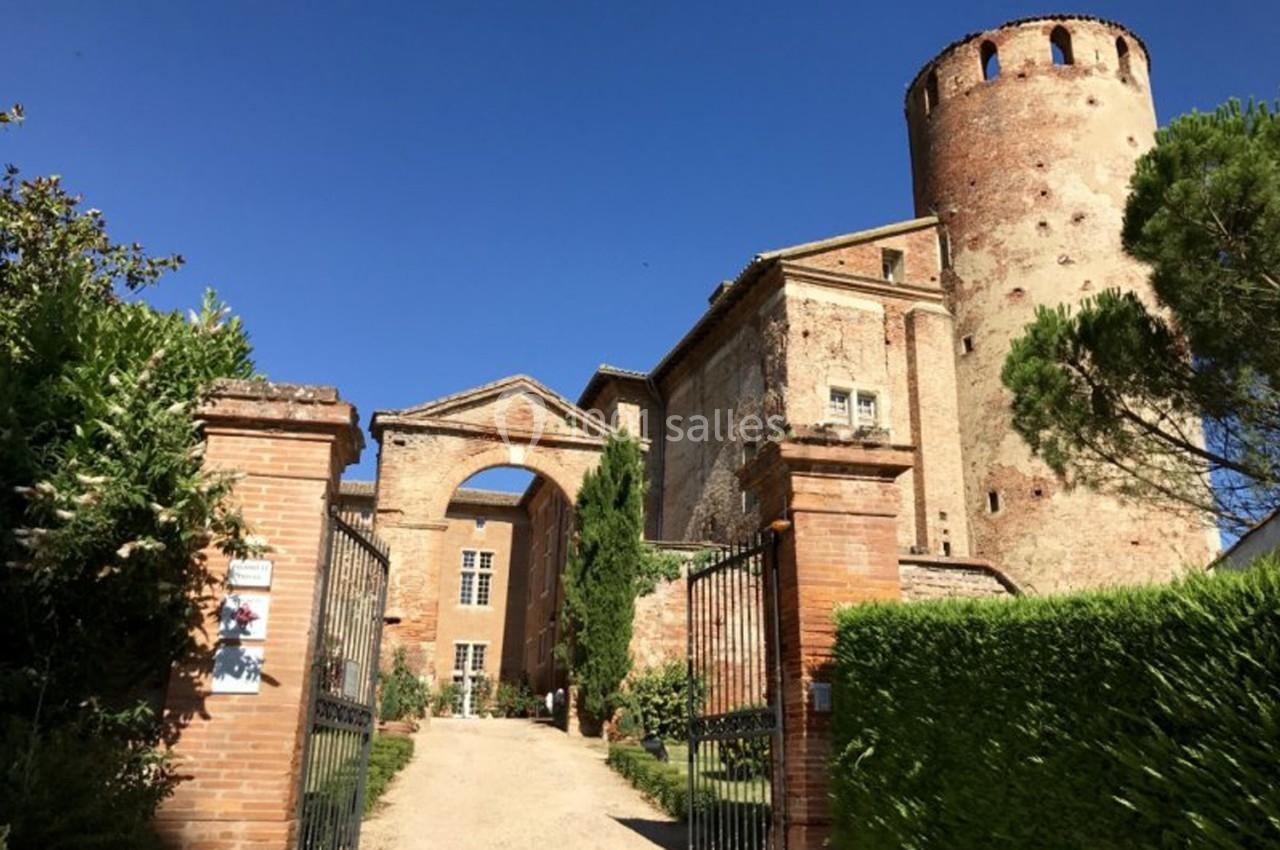 Entrée d'un château en briques avec une tour ronde, un portail ouvert et un chemin bordé de verdure.