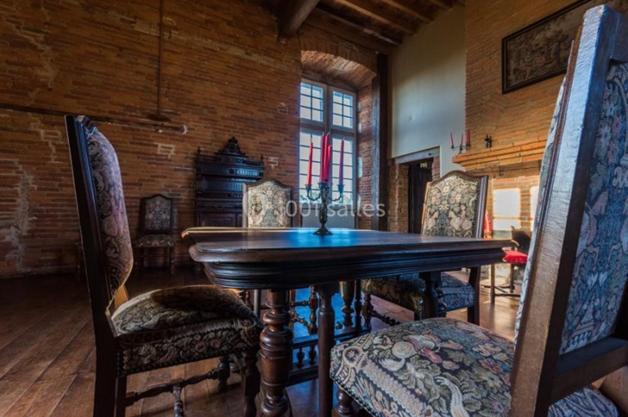 Salle à manger rustique avec table en bois, chaises ornées de motifs et murs en briques, éclairée par une grande fenêtre.