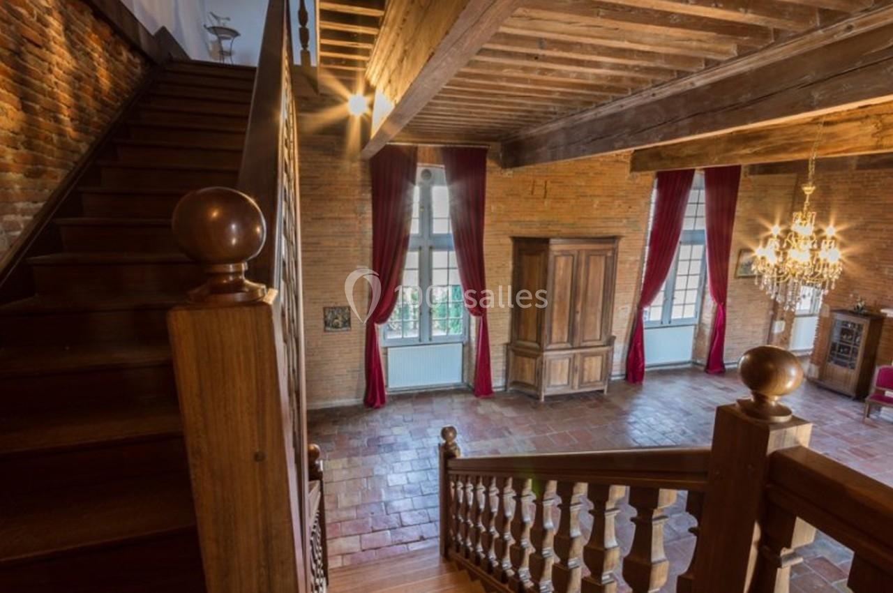 Escalier en bois menant à une pièce lumineuse avec murs en briques, grandes fenêtres, rideaux rouges et lustre.
