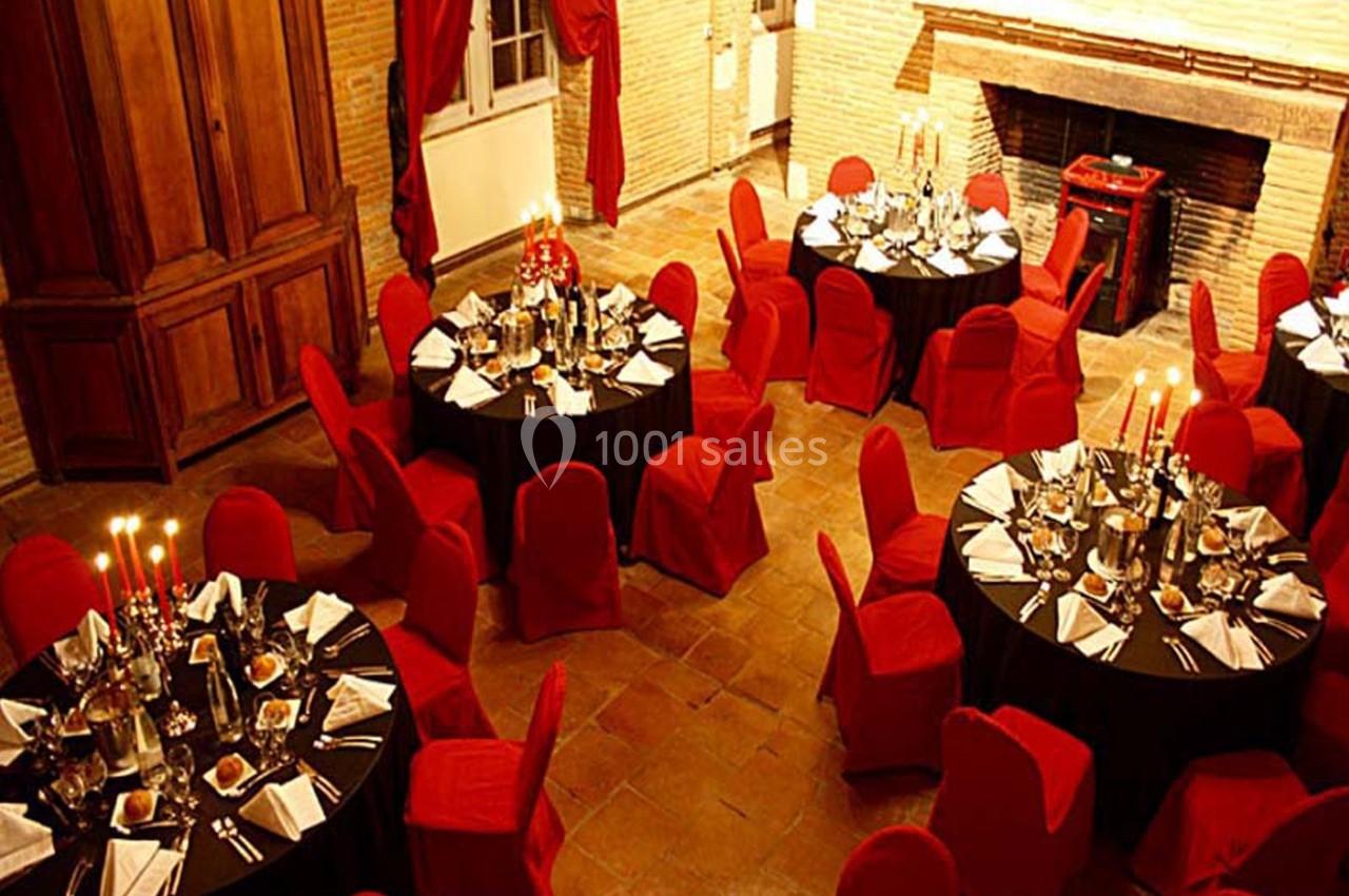 Salle de réception avec tables rondes dressées, nappes noires, chaises rouges et chandeliers allumés.