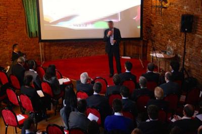 Un conférencier s'adresse à un public assis dans une salle avec un écran de projection et des murs en briques.