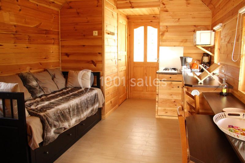Intérieur d'un chalet en bois avec un lit, une kitchenette équipée et une table de travail éclairée par une fenêtre.