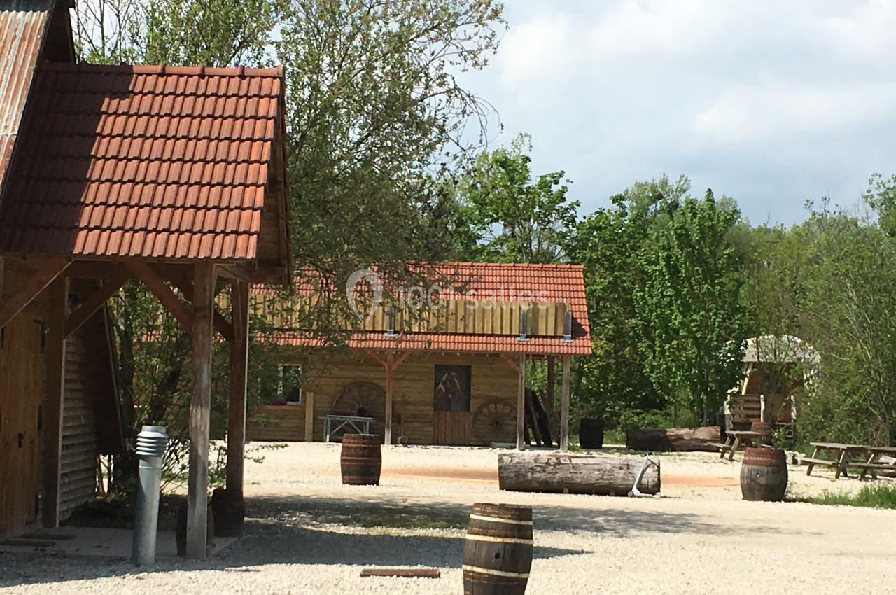 Cour extérieure avec bâtiments en bois, tonneaux, bancs et arbres en arrière-plan sous un ciel partiellement nuageux.