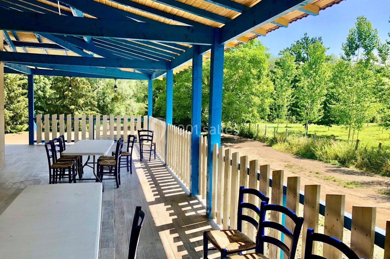 Terrasse couverte avec tables et chaises en bois, entourée d'une barrière, donnant sur un paysage verdoyant.