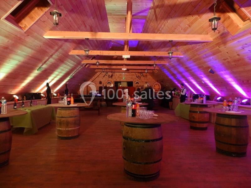 Salle en bois avec plafond en pente, tables en tonneaux et éclairage coloré, préparée pour un événement.