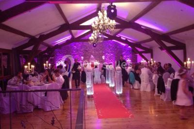 Salle vide avec sol en béton, murs en pierre et bois, tables pliées contre le mur et lustres suspendus au plafond.