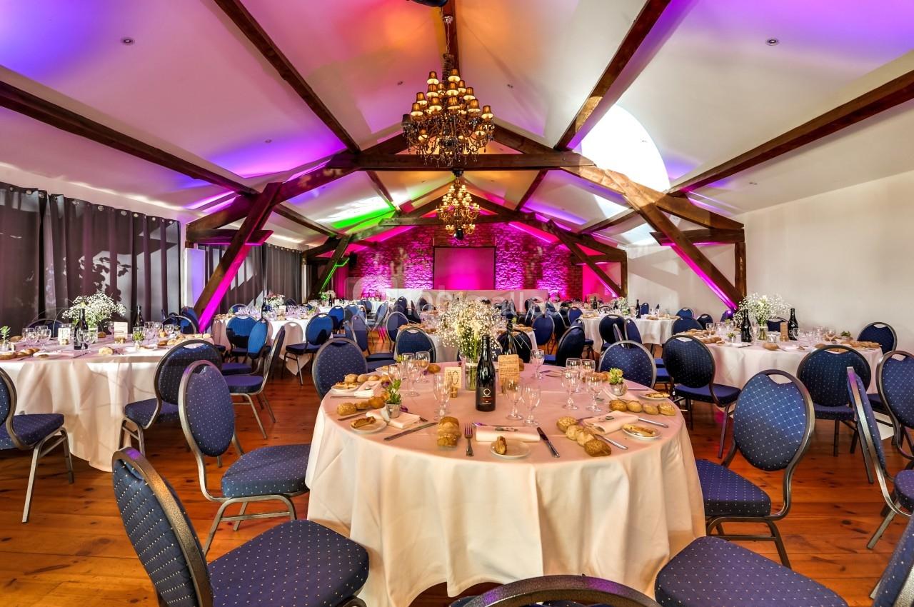 Salle de réception décorée avec des tables rondes dressées, éclairage coloré et poutres apparentes.