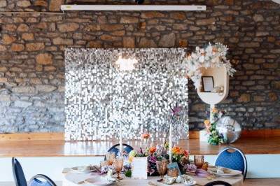 Salle vide avec sol en béton, murs en pierre et bois, tables pliées contre le mur et lustres suspendus au plafond.