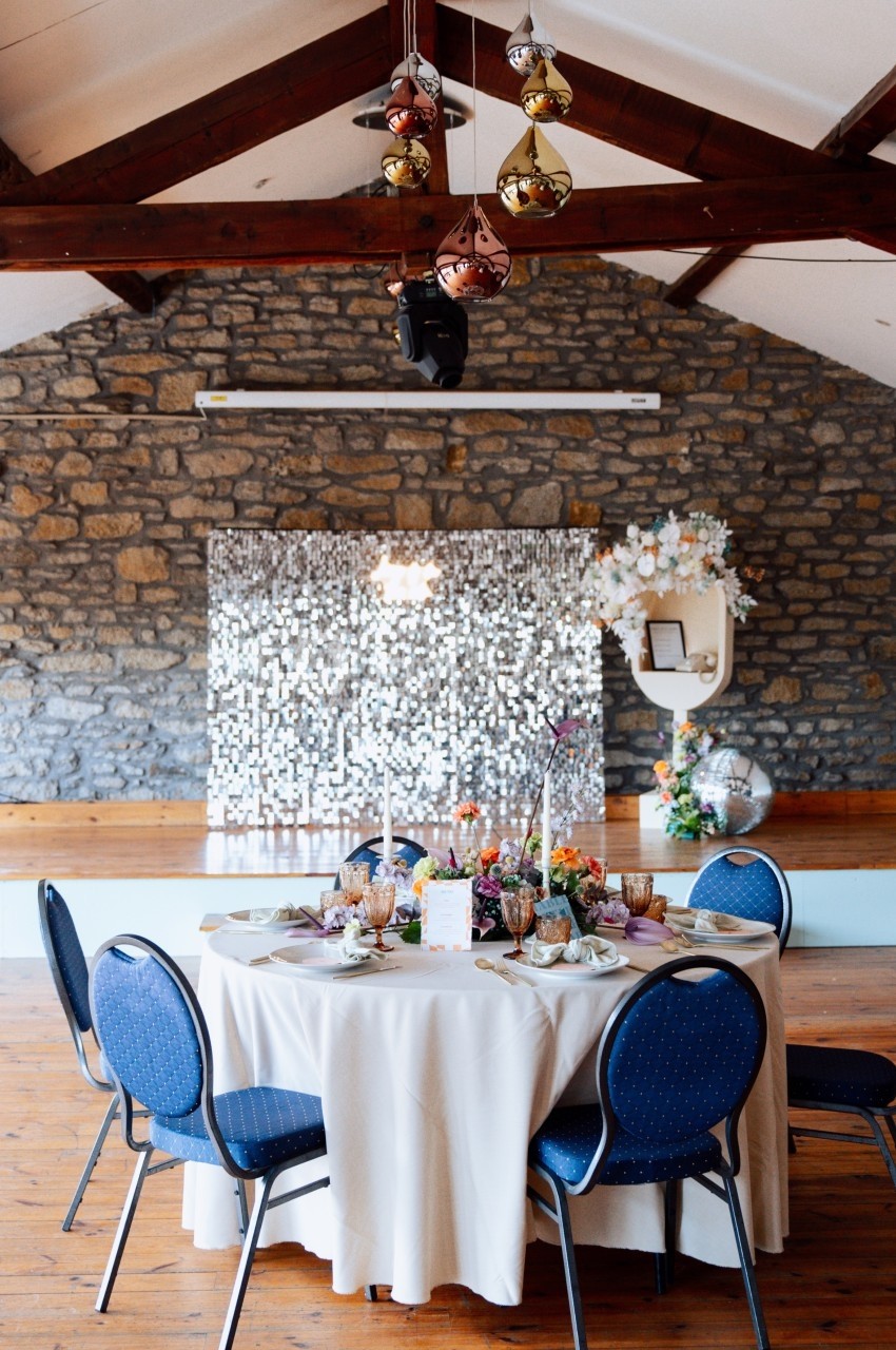 Table ronde décorée avec vaisselle colorée et fleurs, devant un mur en pierre et un fond scintillant dans une salle…