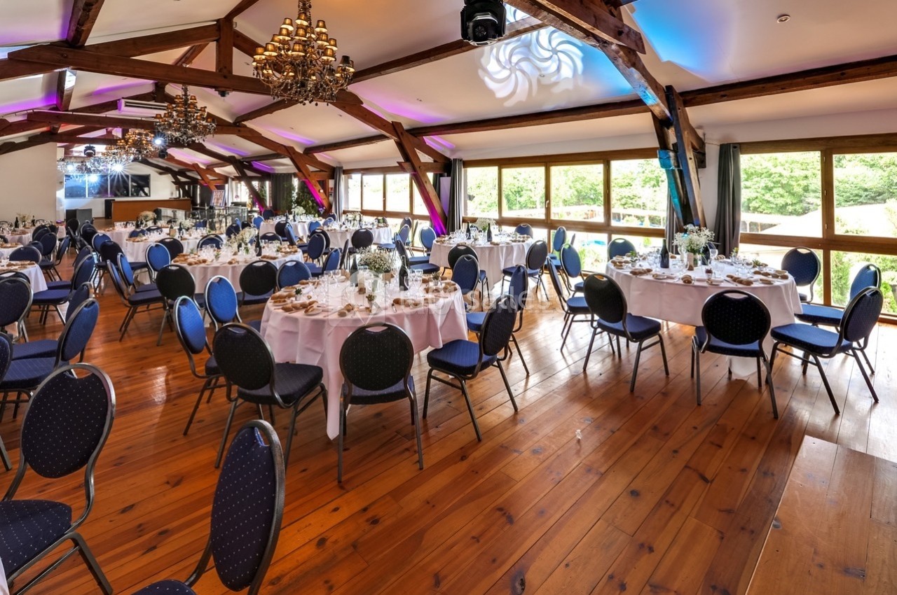 Salle de réception lumineuse avec tables rondes dressées, chaises bleues et poutres apparentes en bois.