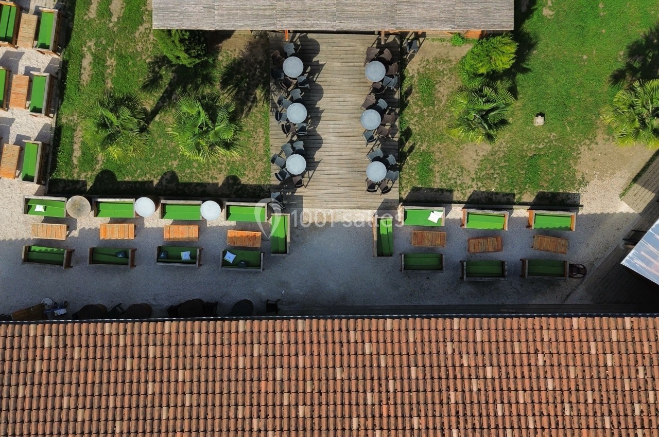 Vue aérienne d'une terrasse avec tables et chaises disposées autour d'allées en bois, entourée de verdure.